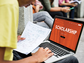 Someone using a laptop which has the word Scholarship displayed on it. The person is holding a piece of paper with formulas on it.