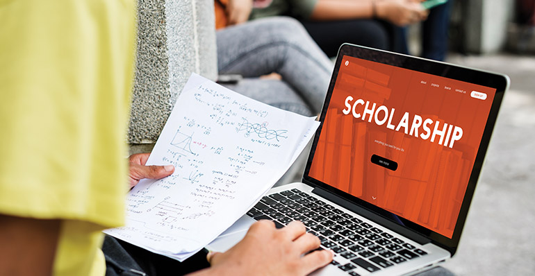 Someone using a laptop which has the word Scholarship displayed on it. The person is holding a piece of paper with formulas on it.